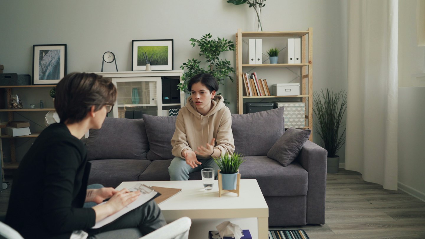 Ein Therapeut mit Klemmbrett hört einem Klienten zu, der auf einem Sofa sitzt.